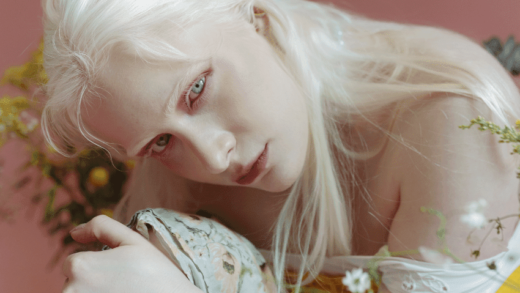 A girl with white hair and skin crouches over her knee holding a flower.