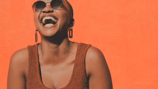 joyful woman with sunglasses and trendy earrings smiling against an orange backdrop