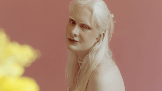 A pale woman with white hair sits looking at the camera. A yellow flower is blurred in the left corner.