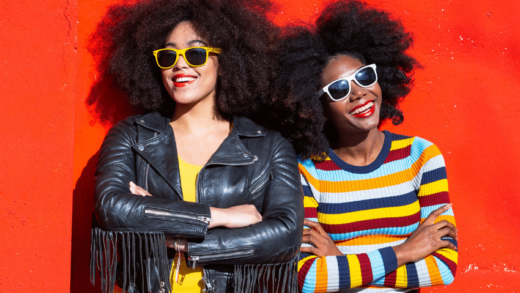 two women with afros in colorful clothing standing against a red backdrop wearing sunglasses smiling boldly