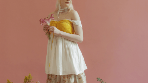 A woman with pale skin and white hair stands amongst fresh cut flowers