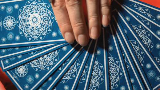 A hand wearing rings holds a fan of blue Tarot cards.